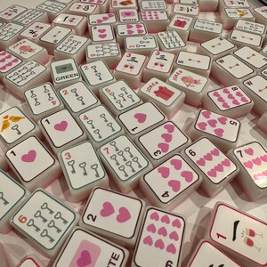 K&ouml;nnte beinhalten: Eine Sammlung wei&szlig;er Mahjong-Steine mit rosa und roten Symbolen, Zahlen und Zeichen. Einige Steine zeigen Herzen, Schl&uuml;ssel und Weingl&auml;ser. Das Wort "WHITE" ist auf einem Stein zu sehen, und "JOKER" auf anderen. Ein Spielset.