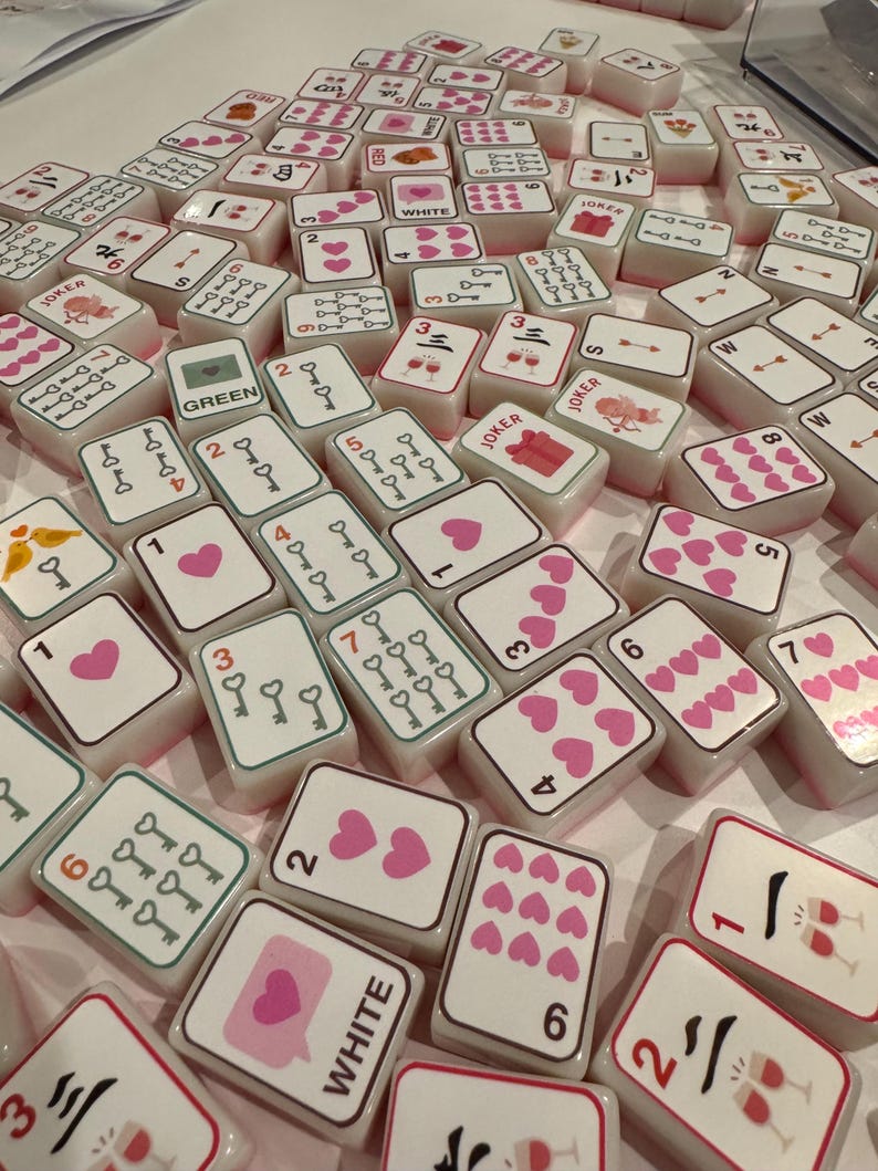 K&ouml;nnte beinhalten: Eine Sammlung wei&szlig;er Mahjong-Steine mit rosa und roten Symbolen, Zahlen und Zeichen. Einige Steine zeigen Herzen, Schl&uuml;ssel und Weingl&auml;ser. Das Wort "WHITE" ist auf einem Stein zu sehen, und "JOKER" auf anderen. Ein Spielset.