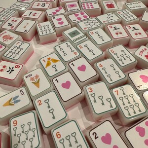 K&ouml;nnte beinhalten: Eine Sammlung wei&szlig;er und rosa Mahjong-Steine mit verschiedenen Symbolen, Zahlen und Anz&uuml;gen. Einige Steine zeigen Herzen, Schl&uuml;ssel und Blumendesigns. Das Wort "JOKER" ist auf einigen Steinen sichtbar.