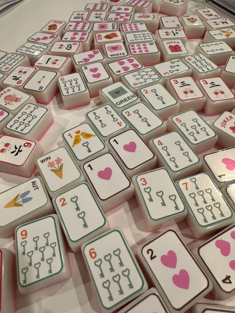 K&ouml;nnte beinhalten: Eine Sammlung wei&szlig;er und rosa Mahjong-Steine mit verschiedenen Symbolen, Zahlen und Anz&uuml;gen. Einige Steine zeigen Herzen, Schl&uuml;ssel und Blumendesigns. Das Wort "JOKER" ist auf einigen Steinen sichtbar.