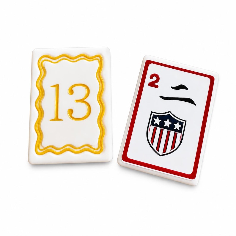 Peut inclure: Deux tuiles de jeu rectangulaires. Une tuile blanche porte le chiffre 13 en or dans un cadre festonn&eacute; dor&eacute;. L'autre tuile a un bord rouge, le chiffre 2 et un &eacute;cusson avec des &eacute;toiles et des rayures.