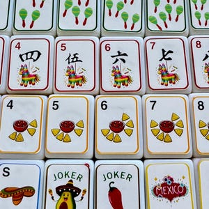 以下が含まれることがあります： 様々なカラフルなイラストが描かれた白いゲームタイル。タイルには数字、マラカス、ピニャータ、サルサとチップス、ソンブレロが描かれています。一部のタイルには「JOKER」と「MEXICO」の文字が書かれています。