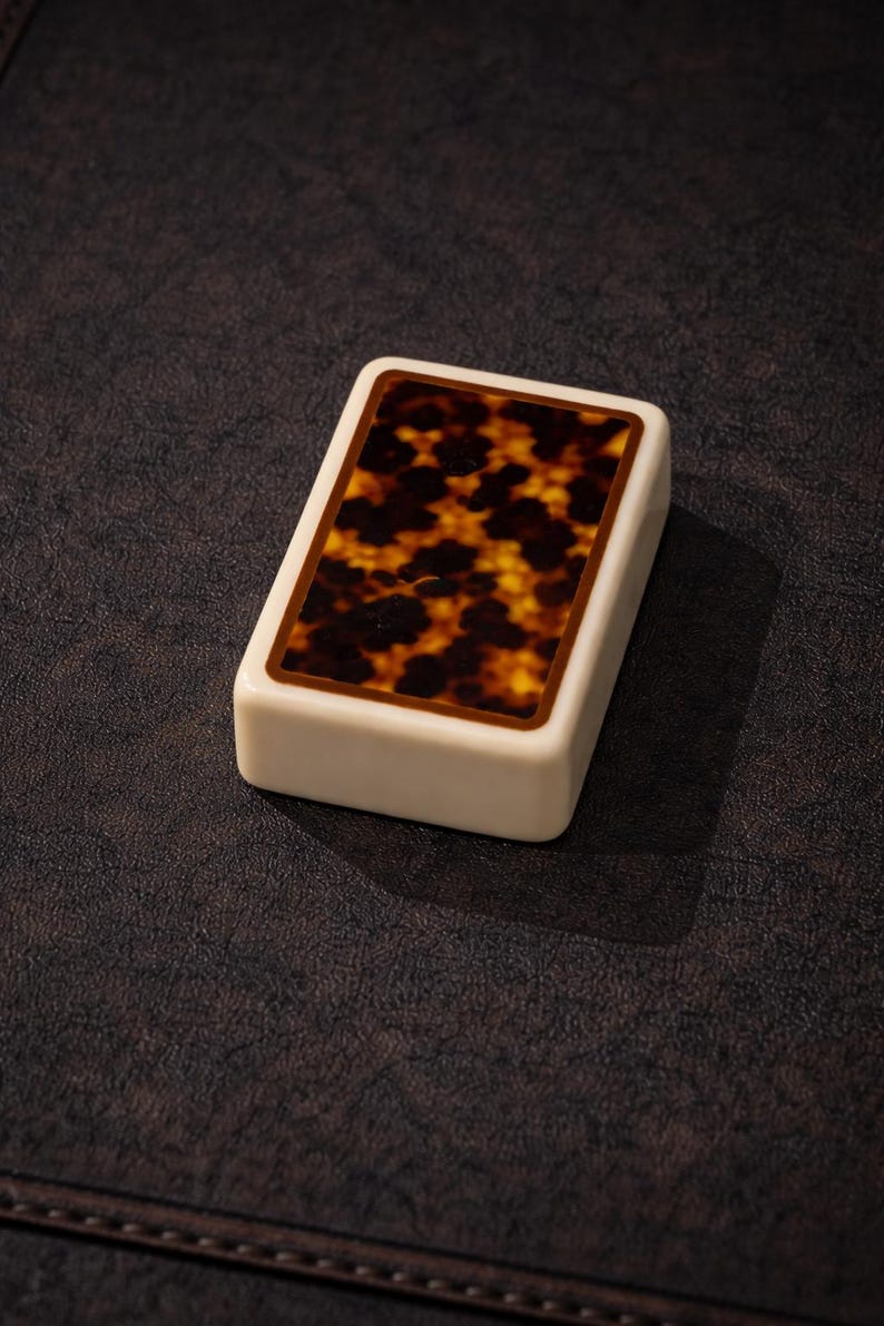 May include: A rectangular, ivory-colored object with a tortoiseshell pattern on the top surface. The tortoiseshell design features a mix of brown and yellow hues, framed by a thin brown border. The object is set against a dark brown background.