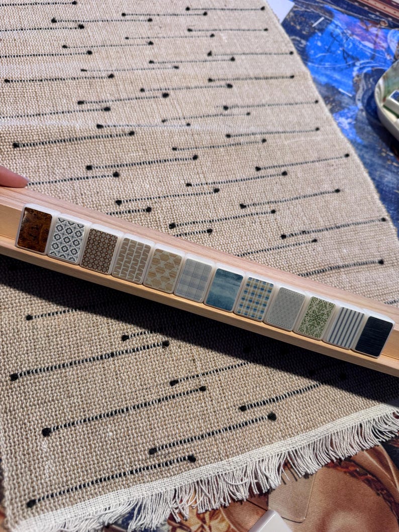 Op de afbeelding: Een houten display met een reeks kleine, patroon tegels. De tegels hebben verschillende ontwerpen, waaronder geometrische patronen, texturen en strepen. De display staat op een beige tapijt met zwarte stippellijnen.