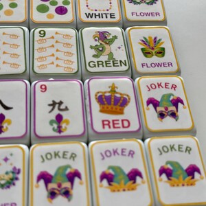 May include: Close-up of white Mahjong tiles with various designs. Some tiles feature the word "WHITE", "GREEN", "RED", and "JOKER". Other tiles display illustrations of flowers, a crown, and a dragon. The tiles have a yellow border.