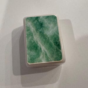 May include: A small, rectangular box with a light-colored frame and a green, marbled surface. The green surface has white veining, creating a natural stone appearance. The box is likely for storing small items.