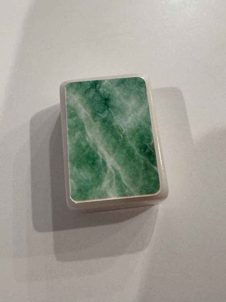 May include: A small, rectangular box with a light-colored frame and a green, marbled surface. The green surface has white veining, creating a natural stone appearance. The box is likely for storing small items.