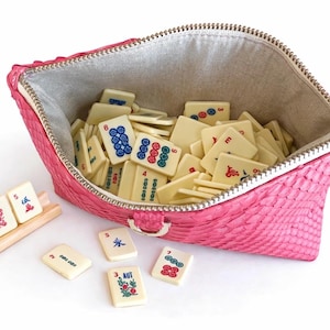 May include: A pink, textured zippered pouch filled with ivory-colored Mahjong tiles. The tiles have various symbols and numbers in red, green, and blue. A wooden rack holds additional tiles, and several loose tiles are scattered around the pouch.