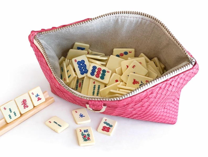 May include: A pink, textured zippered pouch filled with ivory-colored Mahjong tiles. The tiles have various symbols and numbers in red, green, and blue. A wooden rack holds additional tiles, and several loose tiles are scattered around the pouch.
