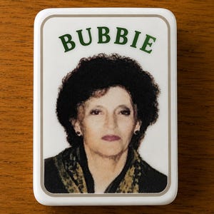 May include: A rectangular white magnet featuring a portrait of a person with dark curly hair and a dark patterned scarf. The word BUBBIE is printed in green capital letters above the portrait, all enclosed within a thin light brown border.