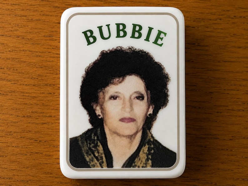 May include: A rectangular white magnet featuring a portrait of a person with dark curly hair and a dark patterned scarf. The word BUBBIE is printed in green capital letters above the portrait, all enclosed within a thin light brown border.