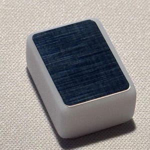 Op de afbeelding: Een rechthoekig, lichtgrijs object met een donkerblauwe, getextureerde bovenkant. De bovenkant heeft een zilveren rand. Het object ligt op een getextureerd, gebroken wit oppervlak.