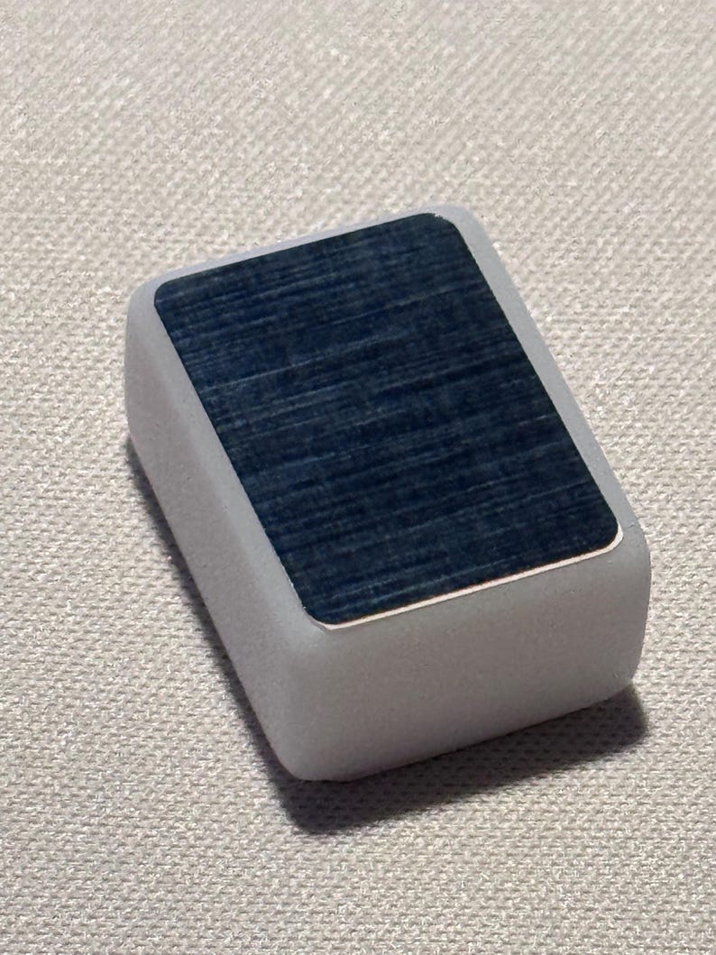 Op de afbeelding: Een rechthoekig, lichtgrijs object met een donkerblauwe, getextureerde bovenkant. De bovenkant heeft een zilveren rand. Het object ligt op een getextureerd, gebroken wit oppervlak.