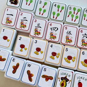 以下が含まれることがあります： カラフルなイラストと数字が描かれた白いゲームタイルのセット。マラカス、ソンブレロ、ピニャータ、サルサやチップスなどの食べ物の画像が特徴です。一部のタイルには中国語の数字と「Joker」の文字があります。