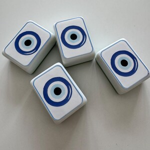 May include: Four white rectangular decorative objects with a blue and black eye design. The eye design features a blue outline, a light blue circle, and a black center. The objects are arranged on a white surface.