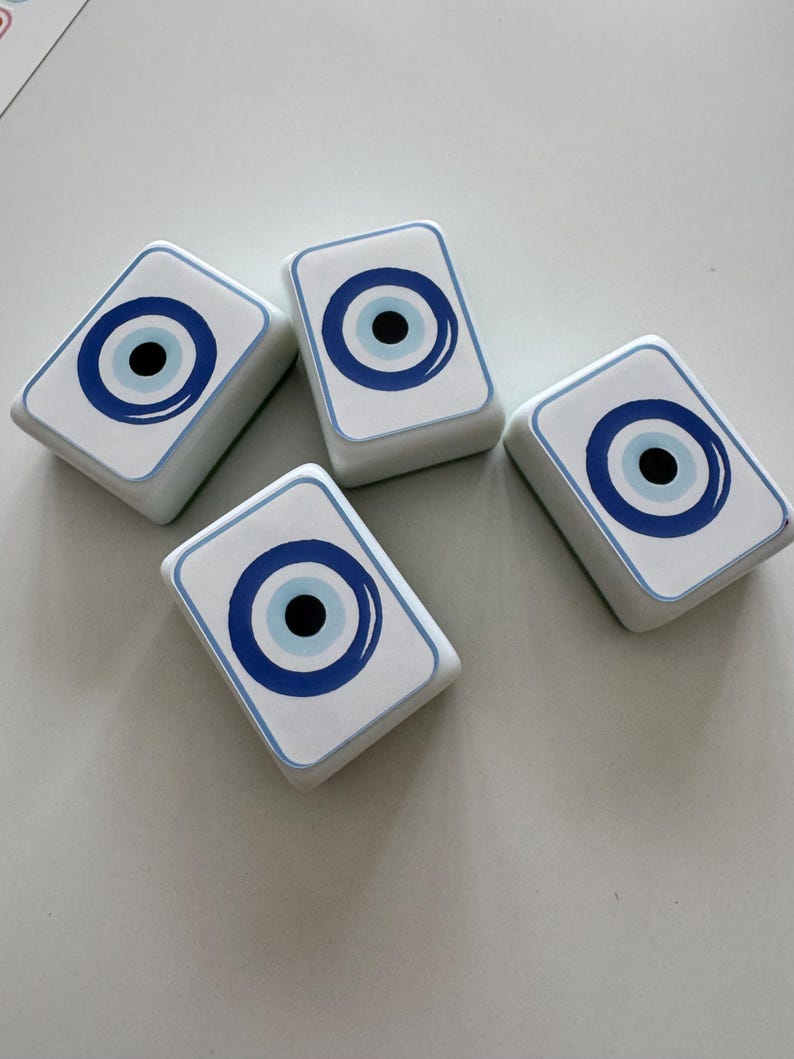 May include: Four white rectangular decorative objects with a blue and black eye design. The eye design features a blue outline, a light blue circle, and a black center. The objects are arranged on a white surface.