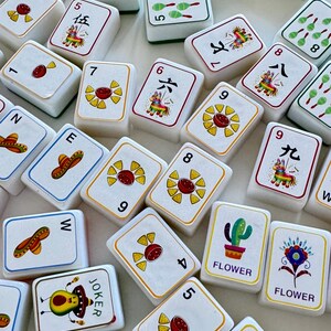 以下が含まれることがあります： カラフルなイラストが描かれた白い麻雀牌のコレクション。牌には数字、シンボル、ピニャータ、ソンブレロ、花の画像が描かれています。牌は明るい表面に散らばっています。