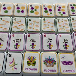 May include: A collection of white Mahjong tiles with various symbols and numbers. The tiles feature dots, trumpets, Chinese characters, and decorative masks with the word "FLOWER". The tiles have purple, green, yellow, and gold accents.