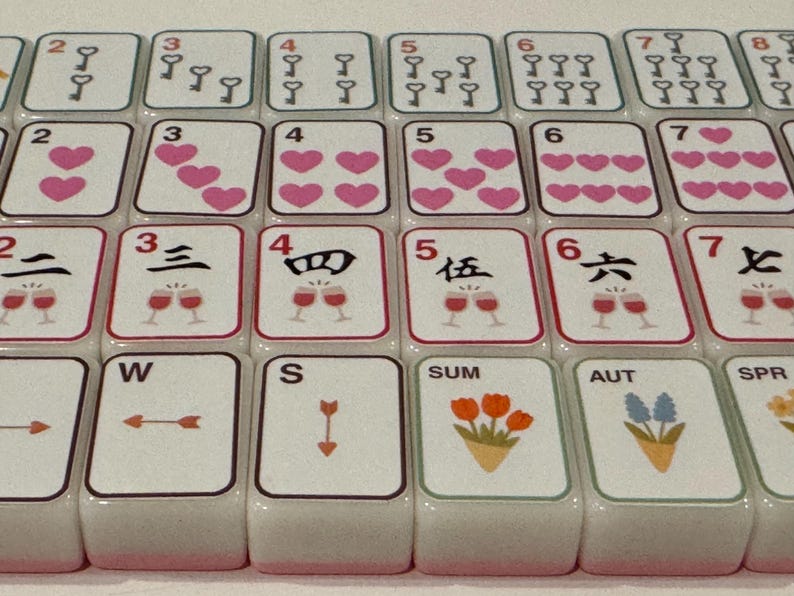 K&ouml;nnte beinhalten: Nahaufnahme eines Mahjong-Spielsets. Die Spielsteine zeigen verschiedene Symbole, darunter Schl&uuml;ssel, Herzen und chinesische Schriftzeichen. Einige Spielsteine zeigen saisonale Bilder wie Blumen. Die Spielsteine sind wei&szlig; mit roten und schwarzen Akzenten.