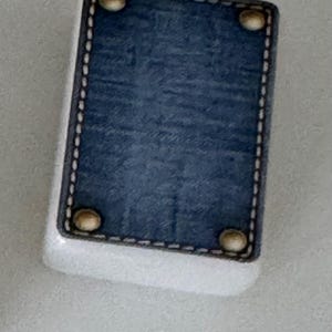 May include: A rectangular, white object with a blue denim-like top. The top is framed with a black border and white stitching. Four gold-colored rivets are placed at each corner of the denim-like top.