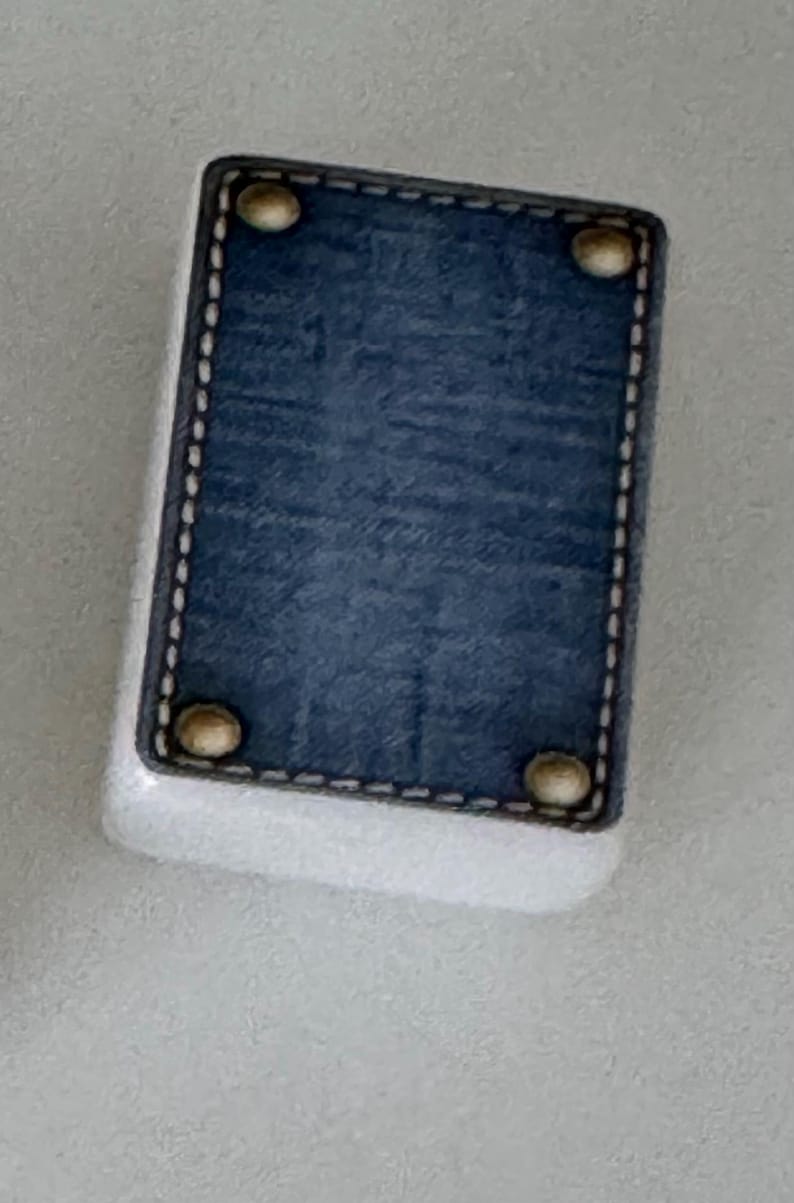May include: A rectangular, white object with a blue denim-like top. The top is framed with a black border and white stitching. Four gold-colored rivets are placed at each corner of the denim-like top.