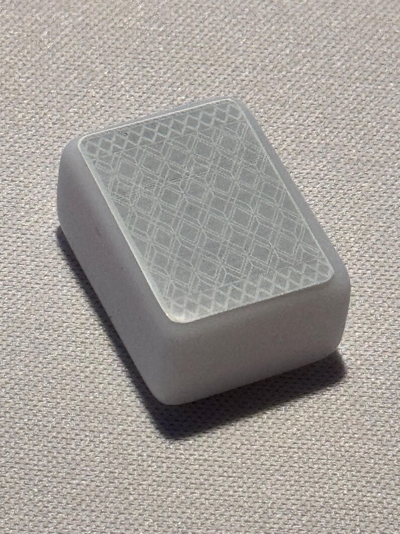Op de afbeelding: Een rechthoekig, grijs object met afgeronde randen. De bovenkant is transparant en heeft een herhalend, ge&euml;tst ruitpatroon. Het is waarschijnlijk een decoratief of functioneel item.