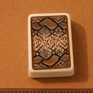 K&ouml;nnte beinhalten: Eine rechteckige Spielkarte mit einem Schlangenmuster in Braun-, Beige- und Schwarzt&ouml;nen. Die Karte hat einen wei&szlig;en Rand und liegt auf einem strukturierten, beigefarbenen Hintergrund. Der untere Teil des Bildes zeigt ein braunes, lederartiges Material.