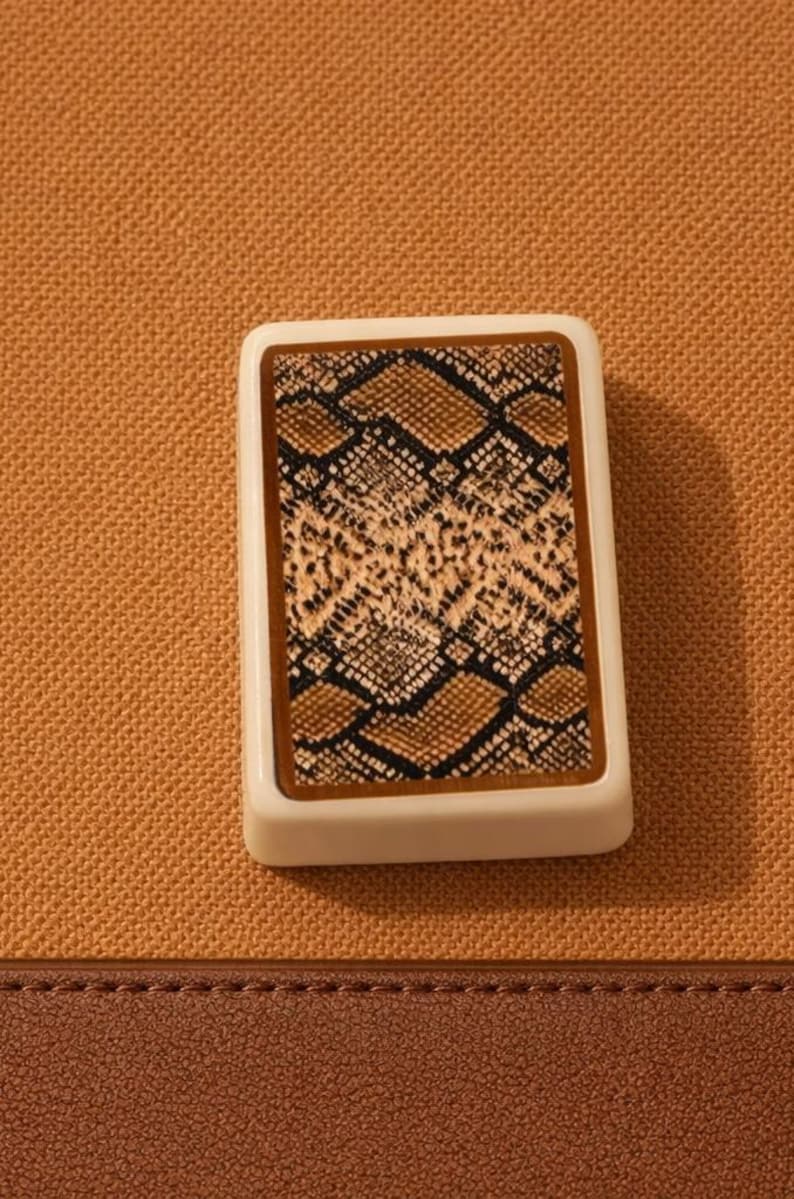 K&ouml;nnte beinhalten: Eine rechteckige Spielkarte mit einem Schlangenmuster in Braun-, Beige- und Schwarzt&ouml;nen. Die Karte hat einen wei&szlig;en Rand und liegt auf einem strukturierten, beigefarbenen Hintergrund. Der untere Teil des Bildes zeigt ein braunes, lederartiges Material.