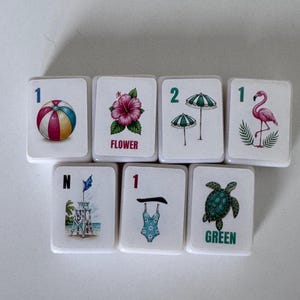 May include: Seven white tiles with colorful summer-themed illustrations. Designs include a beach ball, hibiscus flower with "FLOWER" text, two green umbrellas, a pink flamingo, a lifeguard tower, a blue swimsuit, and a green turtle with "GREEN" text.