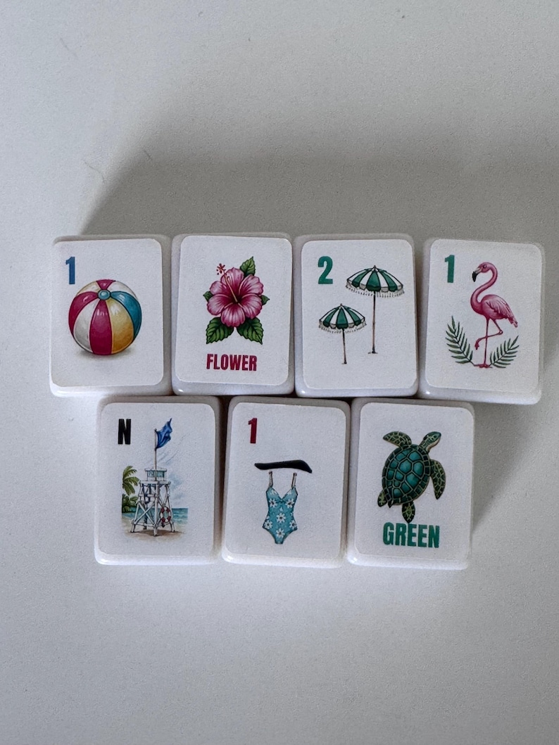 May include: Seven white tiles with colorful summer-themed illustrations. Designs include a beach ball, hibiscus flower with "FLOWER" text, two green umbrellas, a pink flamingo, a lifeguard tower, a blue swimsuit, and a green turtle with "GREEN" text.