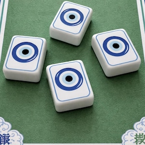 May include: Four white rectangular game tiles with a blue eye design on a green surface. The eye design features a blue outline, a blue iris, and a black pupil. The tiles are arranged on a green surface with a blue border.
