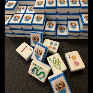 May include: A set of white mahjong tiles with blue edges, featuring custom printed images of a small dog wearing a blue bandana. Some tiles display traditional mahjong symbols, including a green dragon, bamboo, and floral patterns with numbers.