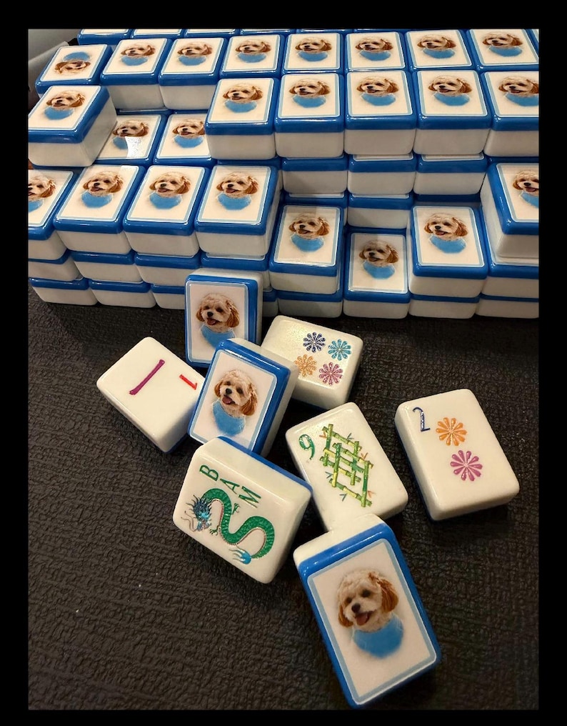 May include: A set of white mahjong tiles with blue edges, featuring custom printed images of a small dog wearing a blue bandana. Some tiles display traditional mahjong symbols, including a green dragon, bamboo, and floral patterns with numbers.
