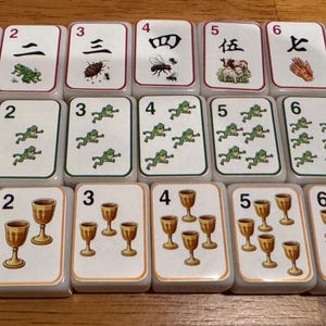May include: A set of rectangular Mahjong tiles with white faces and red borders. The tiles display a variety of symbols, numbers, and illustrations, including Chinese characters, frogs, and goblets. The tiles are arranged in neat rows.