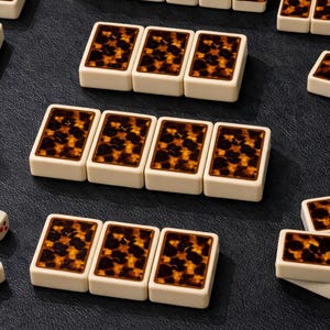 Könnte beinhalten: Eine Ansammlung von elfenbeinfarbenen, rechteckigen Spielsteinen mit einem Schildpattmuster auf der Oberseite. Die Spielsteine sind auf einer dunklen Oberfläche angeordnet, was auf ein Spiel oder Bastelset hindeutet. Das Schildpattmuster zeigt braune und bernsteinfarbene Farbtöne.