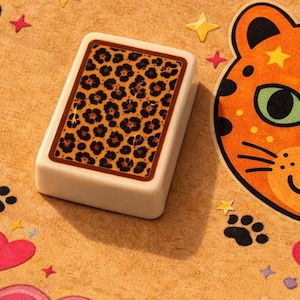 K&ouml;nnte beinhalten: Ein rechteckiges, cremefarbenes Spielteil mit Leopardenmuster. Es liegt auf einer beigefarbenen Oberfl&auml;che, die mit Herzen, Sternen und einem Cartoon-Katzenkopf verziert ist. Der Rand der Oberfl&auml;che ist rosa.