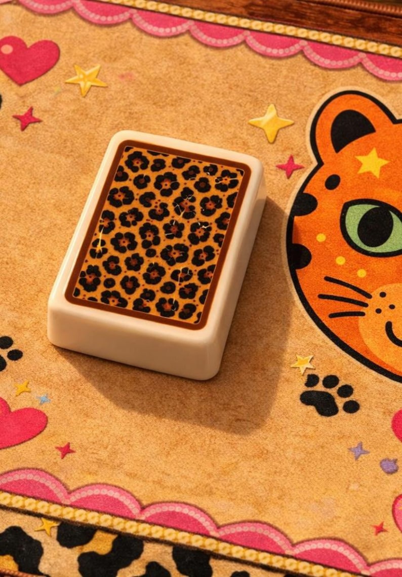 K&ouml;nnte beinhalten: Ein rechteckiges, cremefarbenes Spielteil mit Leopardenmuster. Es liegt auf einer beigefarbenen Oberfl&auml;che, die mit Herzen, Sternen und einem Cartoon-Katzenkopf verziert ist. Der Rand der Oberfl&auml;che ist rosa.