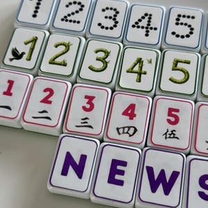 Może przedstawiać: Zbliżenie białych płytek do gry w Mahjong z czarnymi, zielonymi i czerwonymi symbolami i cyframi. Płytki wyświetlają liczby 1-5 w kropkach, bambusie i chińskich cyfrach. Widoczne jest również słowo "NEWS".