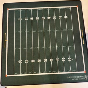 May include: A square green game mat featuring a white American football field gridiron design with yard line markings, numbers, and yellow goal posts. The bottom right corner displays the text FESTIVE MAHJONG BY TARYN ROMY with a small icon.