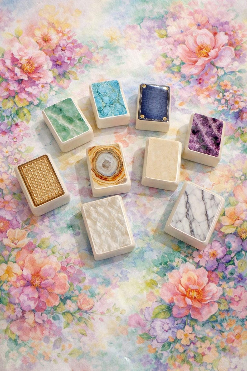 May include: A collection of rectangular decorative objects with various patterned tops, including marble, agate, and textured designs. The objects are arranged on a floral background with pink, purple, and green hues.