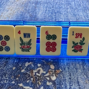 May include: A close-up of ivory-colored Mahjong tiles with various symbols and numbers, including the characters "W" and "五", the number "5" with flower symbols, and the word "WIN". The tiles are arranged in a blue plastic holder.