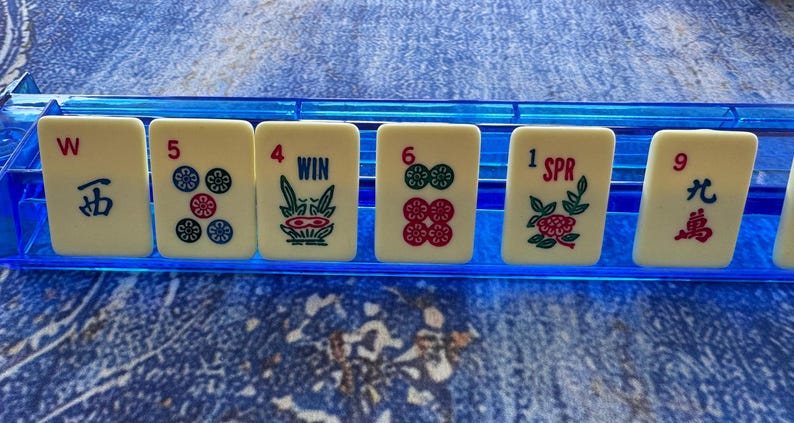May include: A close-up of ivory-colored Mahjong tiles with various symbols and numbers, including the characters "W" and "五", the number "5" with flower symbols, and the word "WIN". The tiles are arranged in a blue plastic holder.