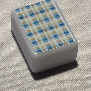 Op de afbeelding: Een klein, rechthoekig, grijs object met een ruitpatroon aan de bovenkant. Het ruitontwerp heeft een mix van blauwe, gele en witte vierkanten. Het object heeft afgeronde randen en een gladde, glanzende afwerking.
