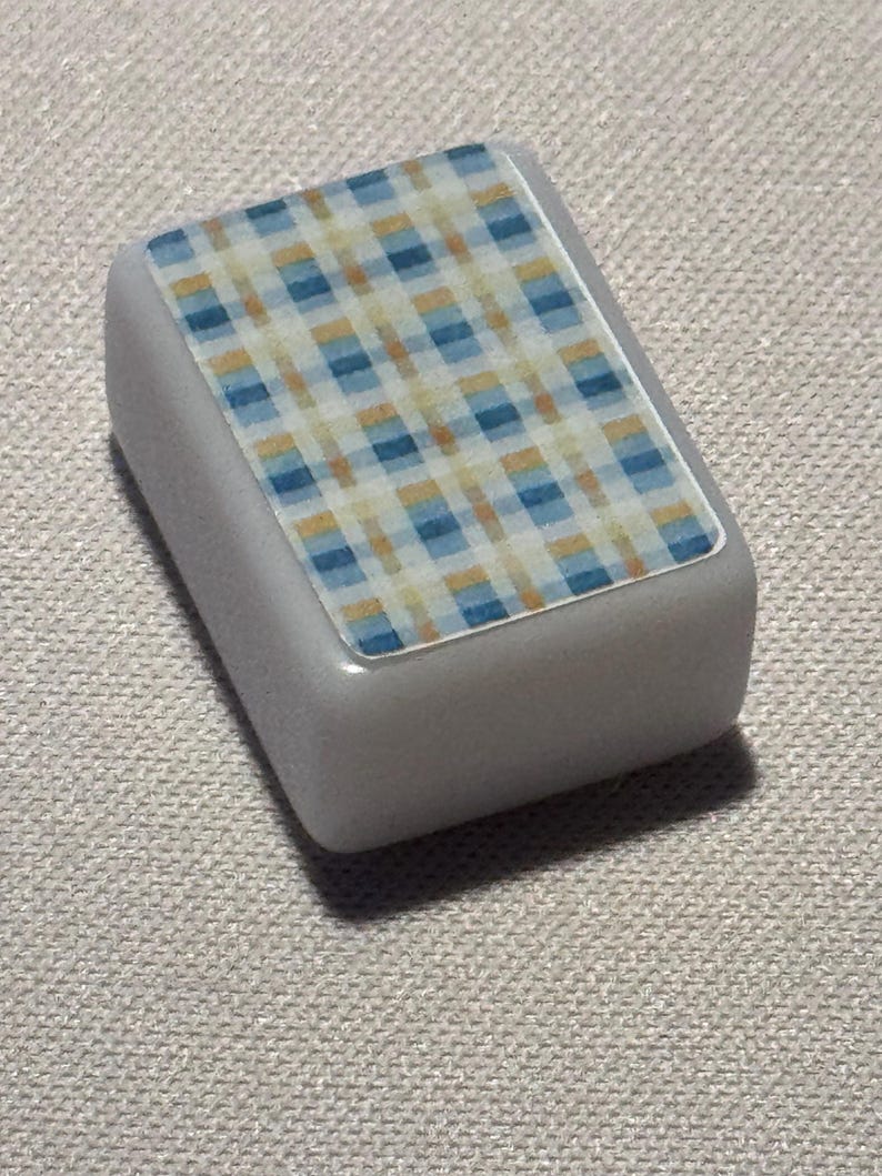 Op de afbeelding: Een klein, rechthoekig, grijs object met een ruitpatroon aan de bovenkant. Het ruitontwerp heeft een mix van blauwe, gele en witte vierkanten. Het object heeft afgeronde randen en een gladde, glanzende afwerking.