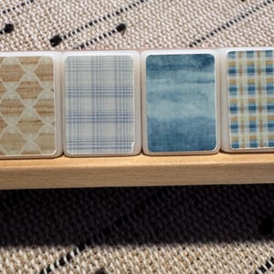 Op de afbeelding: Close-up van een set decoratieve tegels met verschillende patronen. De rechthoekige tegels met afgeronde hoeken zijn geplaatst in een houten bakje. Patronen zijn onder meer een lichtbruin geometrisch ontwerp, blauw geruit, aquarelblauw en een blauw en beige ruitpatroon.