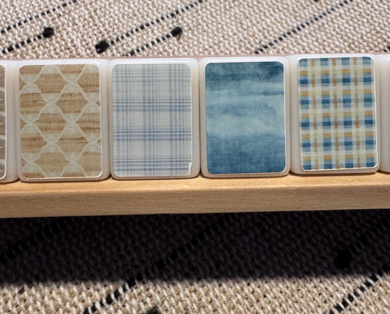 Op de afbeelding: Close-up van een set decoratieve tegels met verschillende patronen. De rechthoekige tegels met afgeronde hoeken zijn geplaatst in een houten bakje. Patronen zijn onder meer een lichtbruin geometrisch ontwerp, blauw geruit, aquarelblauw en een blauw en beige ruitpatroon.