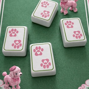 May include: A whimsical game set featuring pink poodles and white tiles with pink paw prints. The tiles have green borders and are arranged on a green surface with pink accents. The poodles wear pink bows and hats.
