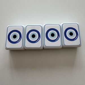 May include: Four white rectangular coasters with a blue and black eye design. Each coaster has a blue border and a central eye motif, creating a symmetrical pattern. The coasters are arranged in a row.