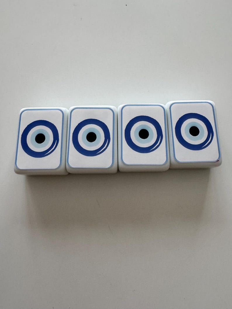 May include: Four white rectangular coasters with a blue and black eye design. Each coaster has a blue border and a central eye motif, creating a symmetrical pattern. The coasters are arranged in a row.