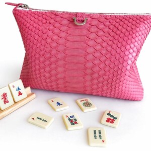 May include: A vibrant pink clutch with a textured snakeskin pattern, a silver zipper, and a small silver ring detail. Several ivory Mahjong tiles with colorful symbols and numbers are scattered around the clutch, and some are in a wooden rack.